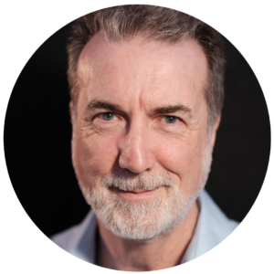 A middle-aged man with grey hair and a beard, wearing a light blue shirt, smiles gently whilst looking at the camera against a dark background—perfect for an Essex web design or creative website design portfolio.