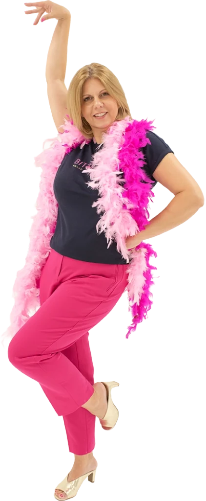 Liz from Bit2flash website design Chelmsford, is posing with one leg up and one arm up in the air. She is wearing bright pink trousers and a navy tshirt with the Bit2flash logo on the front and two pink feather boas around her neck.